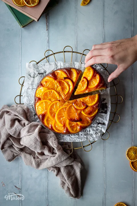 A distanced flat lay of a sticky orange cake with a slice cut out of it on a round cooling rack with a hand reaching in over a cool grey wood effect backdrop. There are also dried orange slices, two stacked books and a light brown linen napkin in the background.