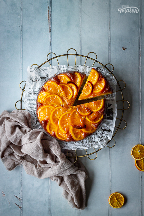 A distanced flat lay of a sticky orange cake with a slice cut out of it on a round cooling rack over a cool grey wood effect backdrop. There are also dried orange slices and a light brown linen napkin in the background.