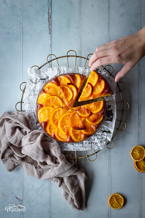 A distanced flat lay of a sticky orange cake with a slice cut out of it on a round cooling rack with a hand reaching in over a cool grey wood effect backdrop. There are also dried orange slices and a light brown linen napkin in the background.