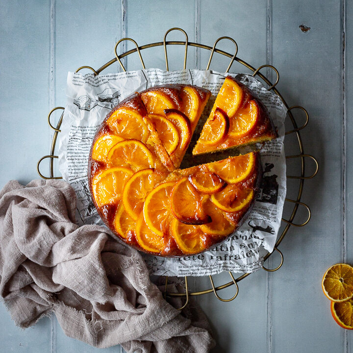 A flat lay of a sticky orange cake with a slice cut out of it on a round cooling rack over a cool grey wood effect backdrop. There are also dried orange slices and a light brown linen napkin in the background.