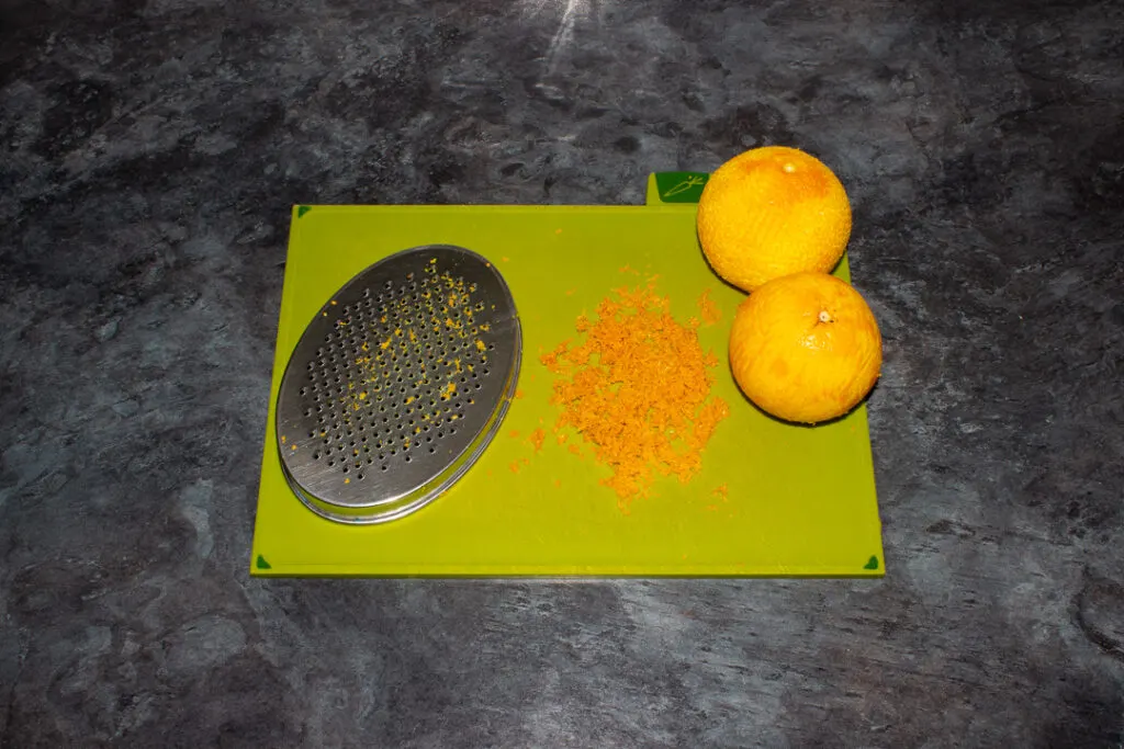 Two zested oranges, the zest and a grater on a green chopping board on a kitchen worktop.