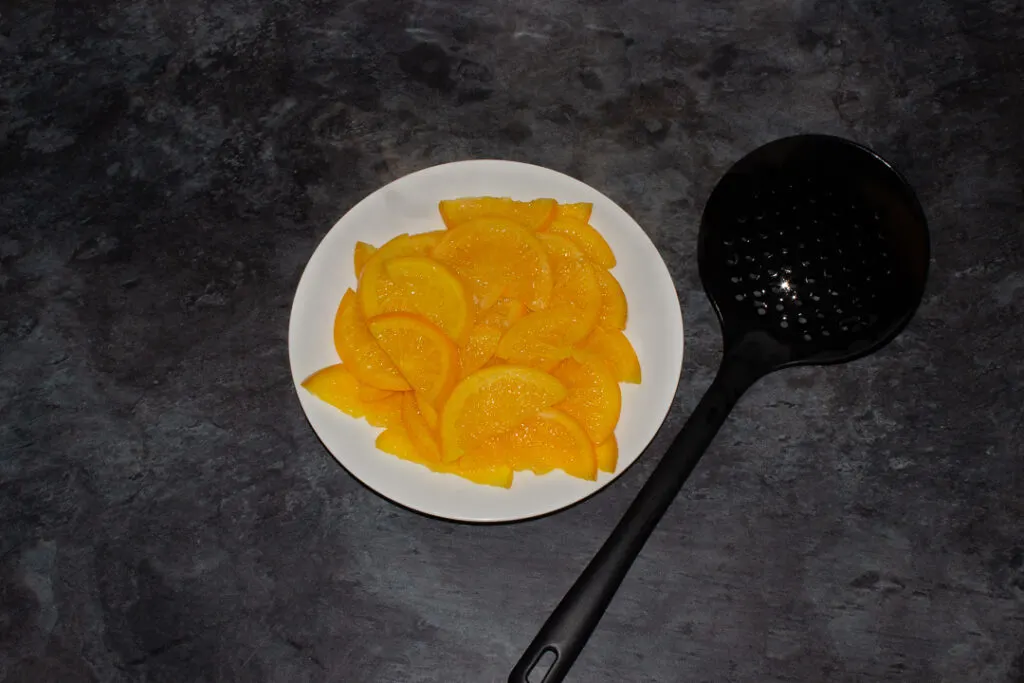 Boiled semicircle orange slices on a white plate with a slotted spoon on a kitchen worktop.