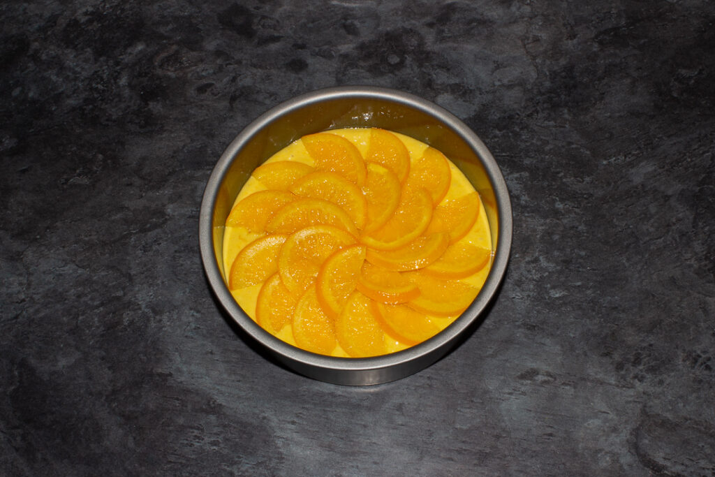 Sticky orange cake batter topped with orange semicircles in a lined round baking tin on a kitchen worktop.