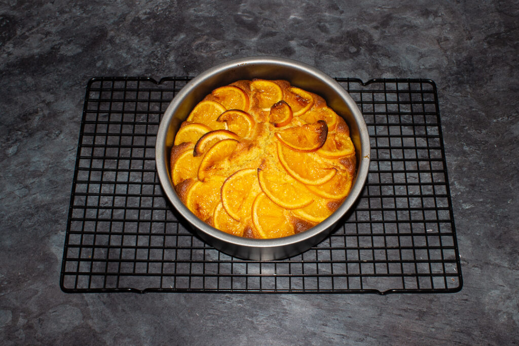 Baked sticky orange cake in a round baking tin on a cooling rack on a kitchen worktop.