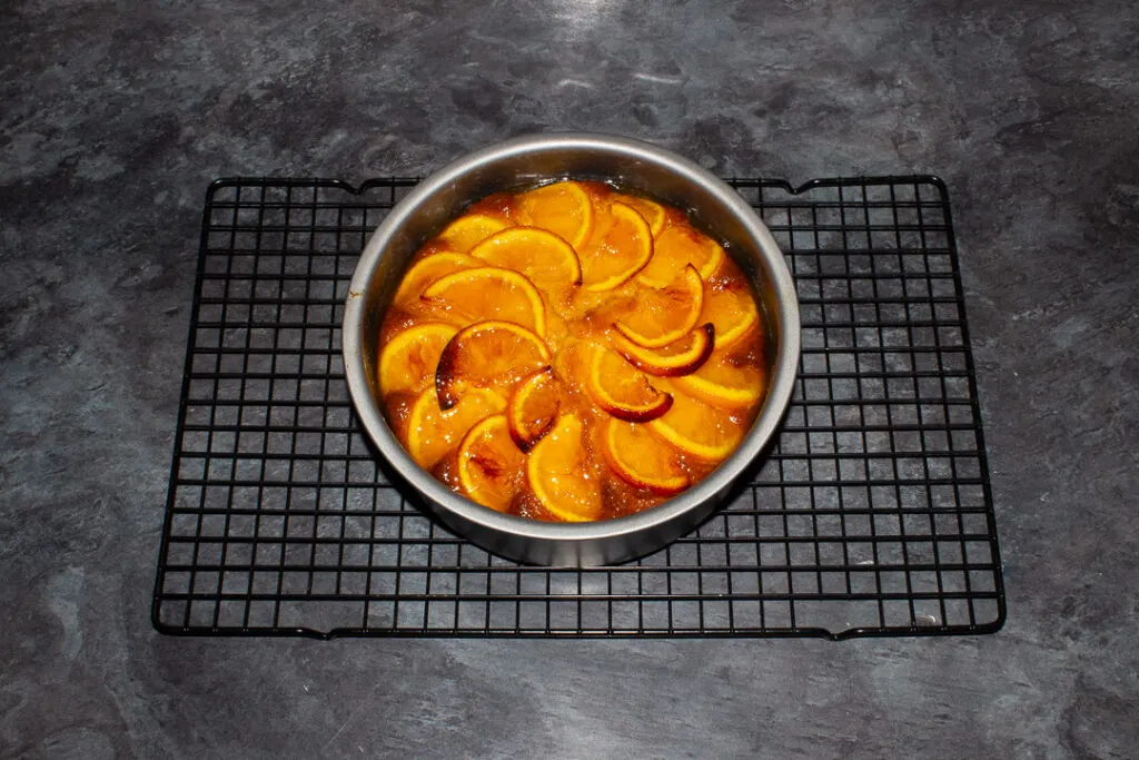 Baked sticky orange cake topped with orange syrup in a round baking tin on a cooling rack on a kitchen worktop.