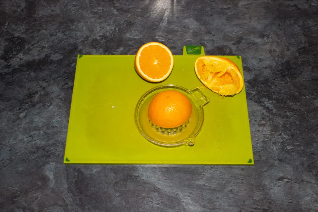 Orange halves being juiced with a glass juicer on a green chopping board on a kitchen worktop.