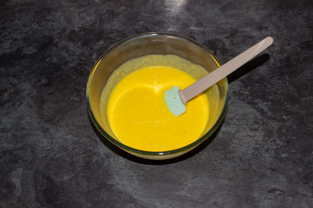 The wet sticky orange cake ingredients mixed together in a glass bowl with a spatula on a kitchen worktop.