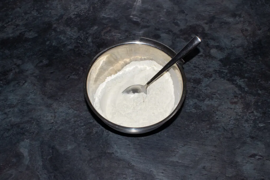 Flour and baking powder in a metal bowl with a metal spoon on a kitchen worktop.
