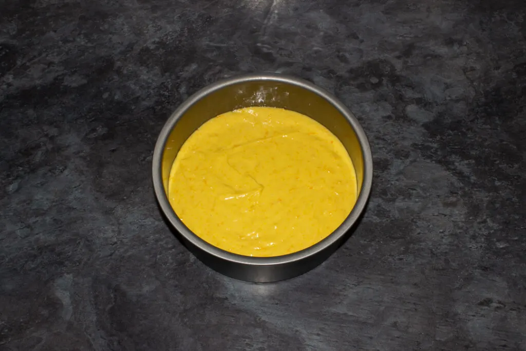 Sticky orange cake batter in a lined round baking tin on a kitchen worktop.