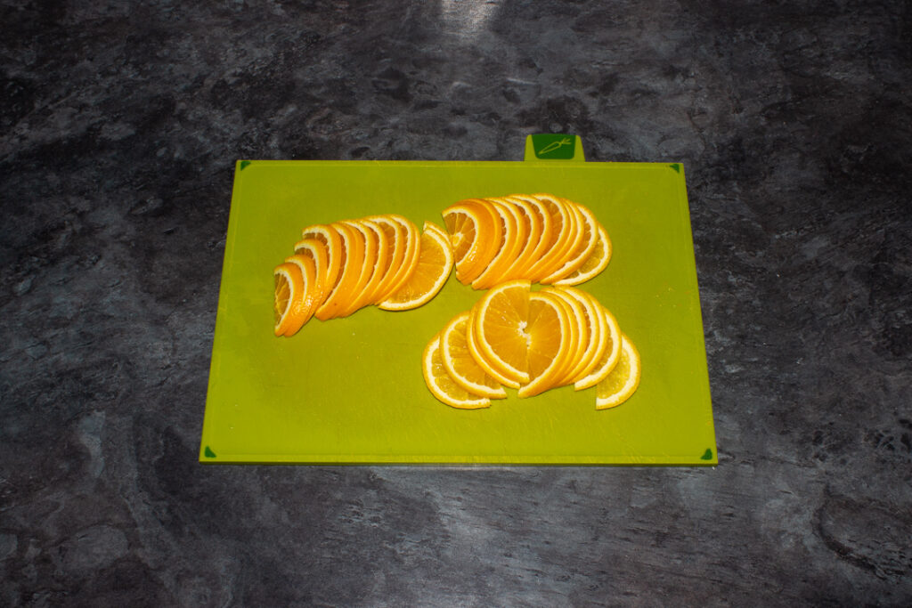 Thinly sliced semicircle orange slices on a green chopping board on a kitchen worktop.