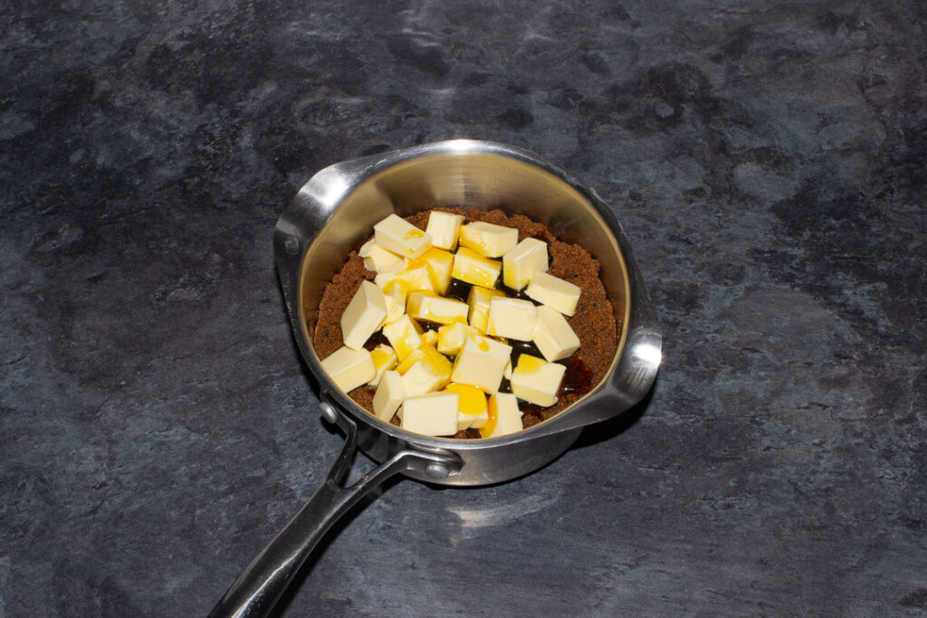 A small saucepan filled with dark muscovado sugar, cubed unsalted butter and golden syrup on a kitchen worktop.