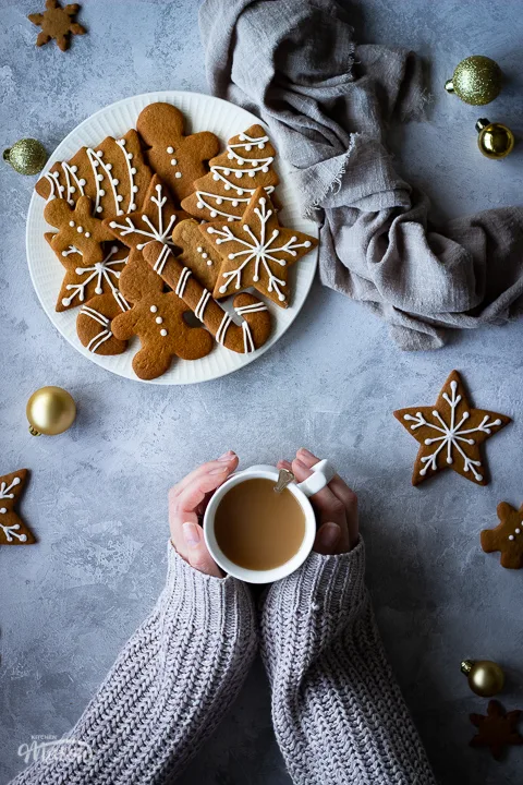 Hands holding a cup of tea next to a white plate filled with decorated gingerbread biscuits with a light brown linen napkin scrunched up to the side. There's also more biscuits and gold baubles scattered in the background. All set over a grey plaster effect backdrop.