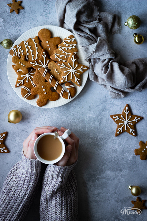 Hands holding a cup of tea off to the side, next to a white plate filled with decorated gingerbread biscuits with a light brown linen napkin scrunched up to the side. There's also more biscuits and gold baubles scattered in the background. All set over a grey plaster effect backdrop.