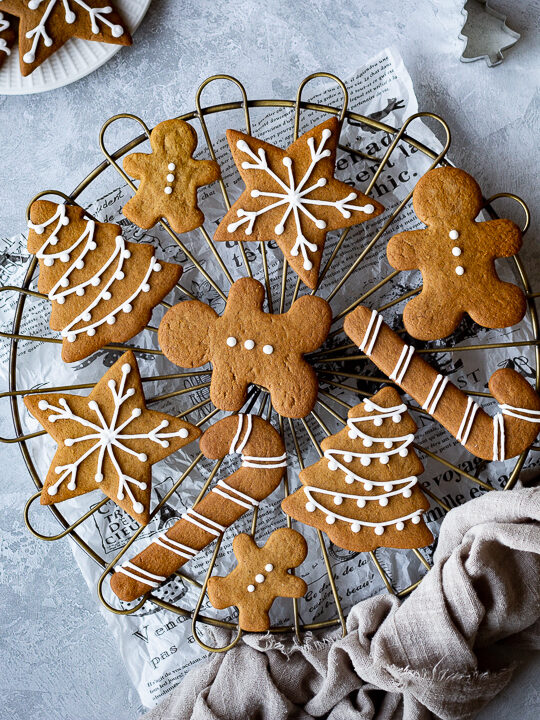 A cooling rack topped with iced gingerbread biscuits set over a scrunched up piece of white baking paper. In the background there is a light brown linen napkin, more iced gingerbread biscuits, some gold baubles, cookie cutters and a white plate with more biscuits on it. All set on a grey plaster effect backdrop.