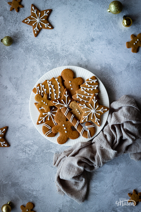 A white plate filled with gingerbread biscuits with a light brown linen napkin scrunched up to the side. There are more biscuits and gold baubles scattered around and it's all set on a grey plaster effect backdrop.