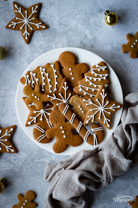 A close up of a white plate filled with gingerbread biscuits with a light brown linen napkin scrunched up to the side. There are more biscuits and gold baubles scattered around and it's all set on a grey plaster effect backdrop.