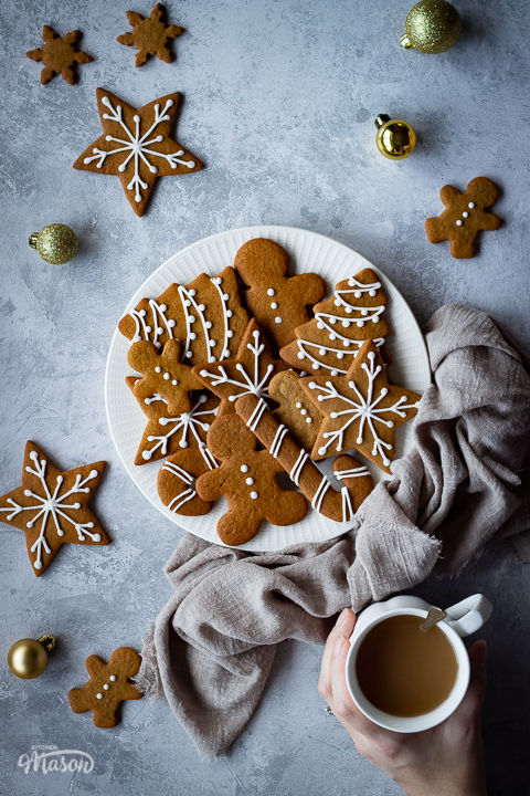 A white plate filled with gingerbread biscuits with a light brown linen napkin scrunched up to the side. There are more biscuits, gold baubles and a hand holding a cup of tea in the background and it's all set on a grey plaster effect backdrop.