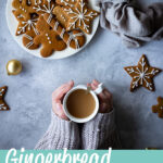 Hands holding a cup of tea off to the side, next to a white plate filled with decorated gingerbread biscuits with a light brown linen napkin scrunched up to the side. There's also more biscuits and gold baubles scattered in the background. All set over a grey plaster effect backdrop. A text overlay says "gingerbread biscuits recipe".