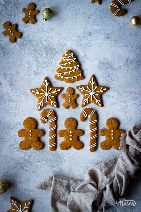 9 decorated gingerbread biscuits laid on a grey plaster effect backdrop in a triangle shape with a light brown linen napkin scrunched up to the side. There are more biscuits and gold baubles in the background.