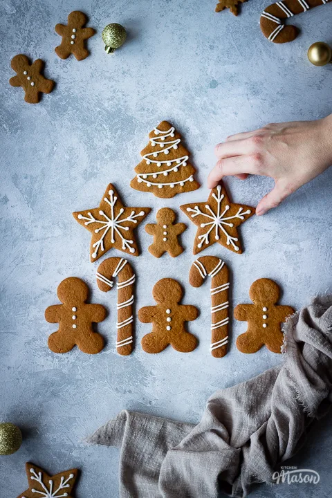 A hand reaching in to 1 of 9 decorated gingerbread biscuits laid on a grey plaster effect backdrop in a triangle shape with a light brown linen napkin scrunched up to the side. There are more biscuits and gold baubles in the background.