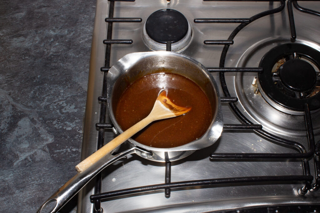 A small saucepan filled with a melted mixture of dark muscovado sugar, unsalted butter and golden syrup set on a stove top.
