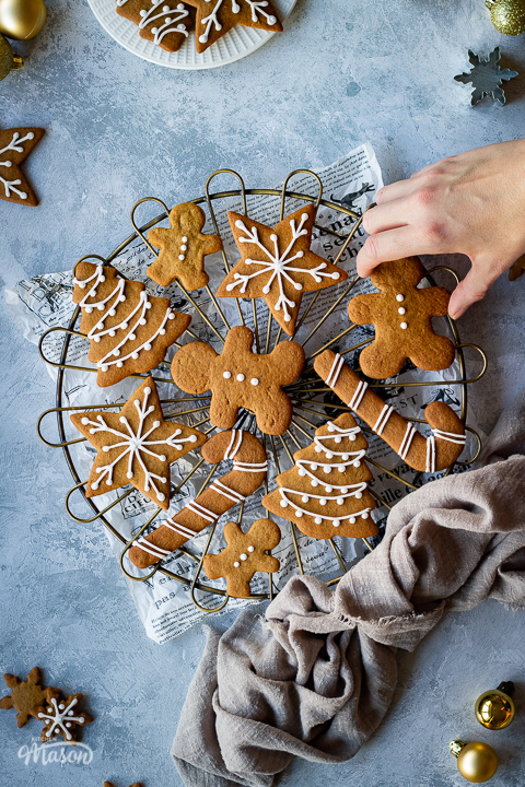 A hand reaching into a cooling rack topped with iced gingerbread biscuits set over a scrunched up piece of white baking paper. In the background there is a light brown linen napkin, more iced gingerbread biscuits, some gold baubles, cookie cutters and a white plate with more biscuits on it. All set on a grey plaster effect backdrop.