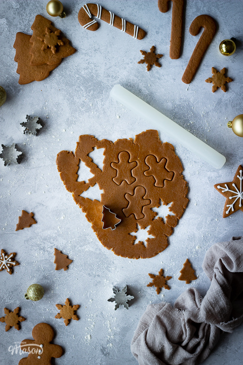 Gingerbread biscuit dough rolled out over a grey plaster effect backdrop with Christmas shapes cut out of it and a cutter on top. In the background there is a small white rolling pin, cut out biscuit shapes, cutters, decorated gingerbread biscuits, gold baubles and a light brown linen napkin.