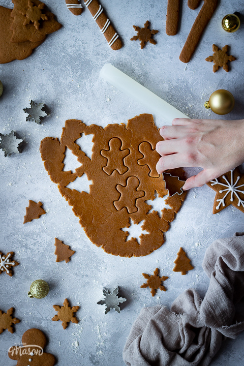 A hand reaching in to some gingerbread biscuit dough rolled out over a grey plaster effect backdrop with Christmas shapes cut out of it and a cutter on top. In the background there is a small white rolling pin, cut out biscuit shapes, cutters, decorated gingerbread biscuits, gold baubles and a light brown linen napkin.