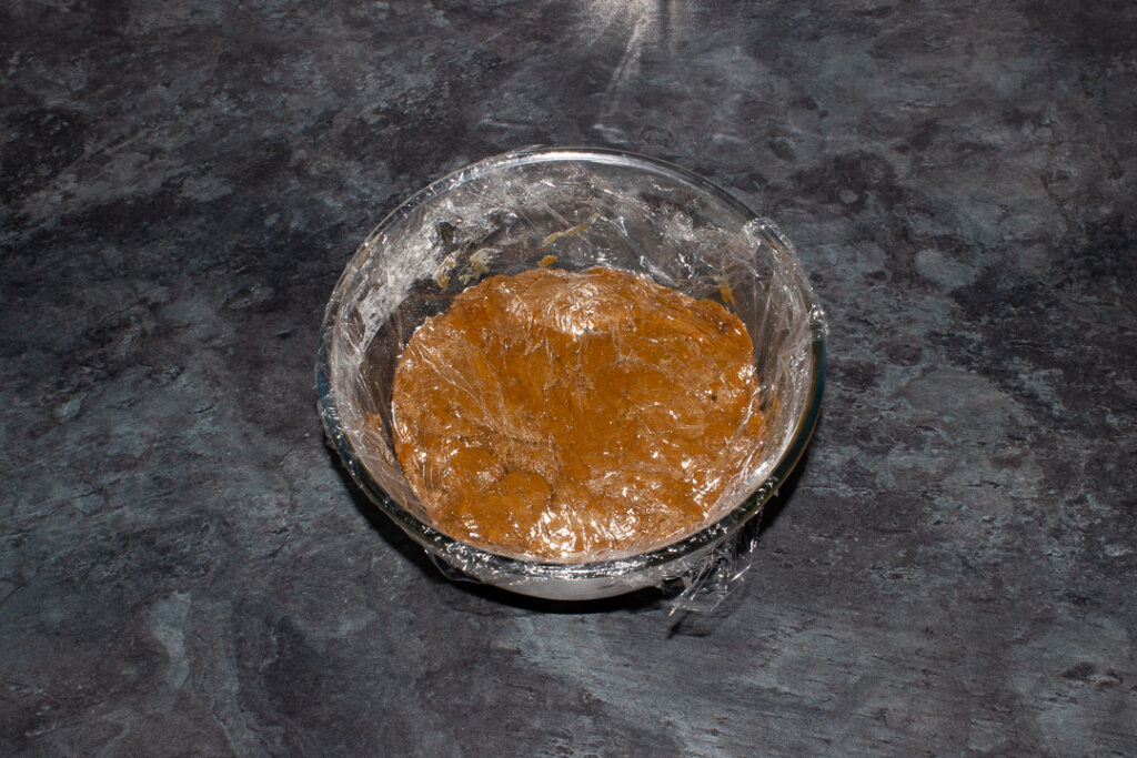 Clingfilm covered gingerbread biscuit dough in a large bowl on a kitchen worktop.