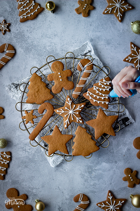 Gingerbread biscuits being decorated on a cooling rack set over a piece of scrunched up baking paper over a grey plaster effect backdrop. There are gold baubles and decorated gingerbread biscuits around the edge of the image.