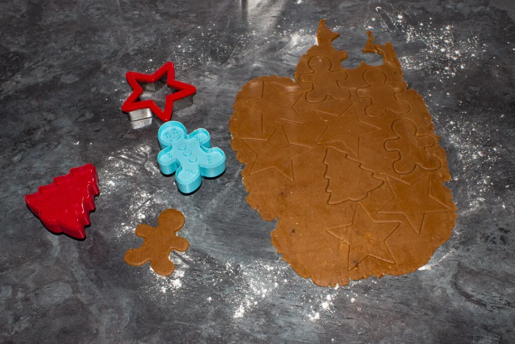 Rolled out gingerbread biscuits that have been cut with Christmas shaped cookie cutters on a kitchen worktop.