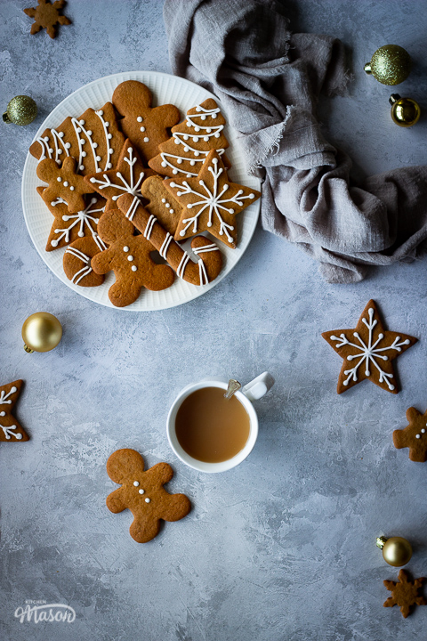 A white plate filled with decorated gingerbread biscuits with a light brown linen napkin scrunched up to the side. There's also a cup of tea, more biscuits and gold baubles scattered in the background. All set over a grey plaster effect backdrop.