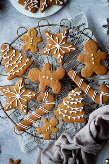 A cooling rack topped with iced gingerbread biscuits set over a scrunched up piece of white baking paper. In the background there is a light brown linen napkin, more iced gingerbread biscuits, some gold baubles, cookie cutters and a white plate with more biscuits on it. All set on a grey plaster effect backdrop.