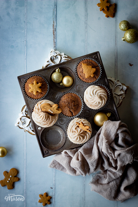 A top down view of iced and none iced gingerbread cupcakes in a muffin pan on a decorative white board. There's some mini gingerbread biscuits, gold baubles and a light brown linen napkin in the background. Set on a cool grey wood effect backdrop.