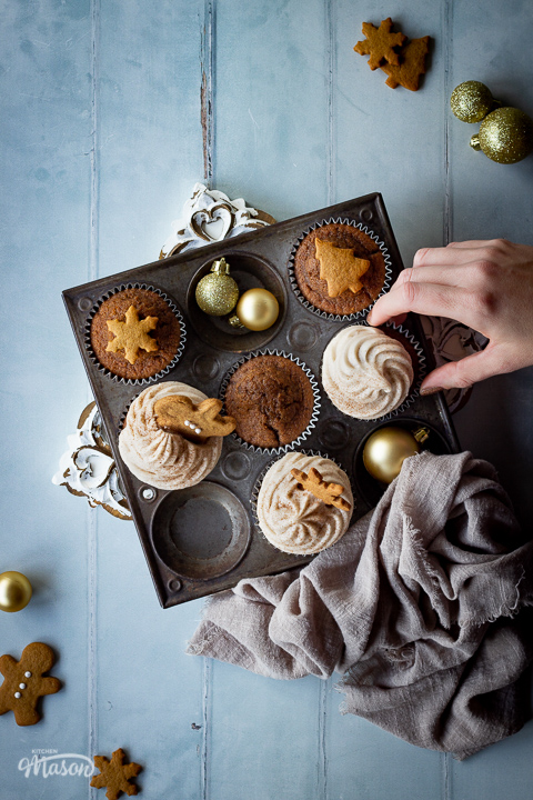 A top down view of a hand reaching in to some iced and none iced gingerbread cupcakes in a muffin pan on a decorative white board. There's some mini gingerbread biscuits, gold baubles and a light brown linen napkin in the background. Set on a cool grey wood effect backdrop.