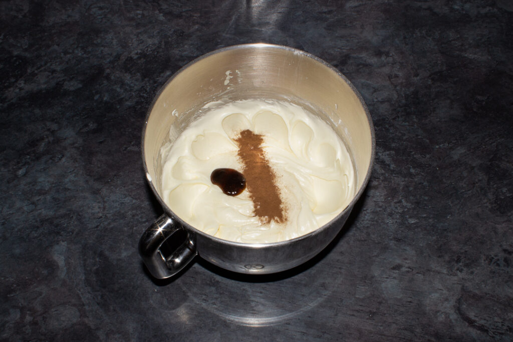 Cream cheese frosting and vanilla/cinnamon in a large silver bowl with a spatula. Set on a kitchen worktop.