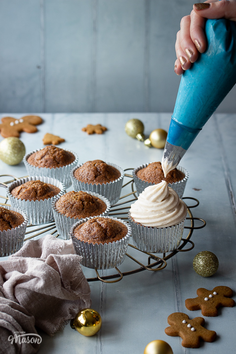A round cooling rack topped with gingerbread cupcakes, one is being iced with cream cheese frosting. There are mini gingerbread biscuits, gold baubles and a light brown linen napkin in the background. Set on a cool grey wood effect backdrop.