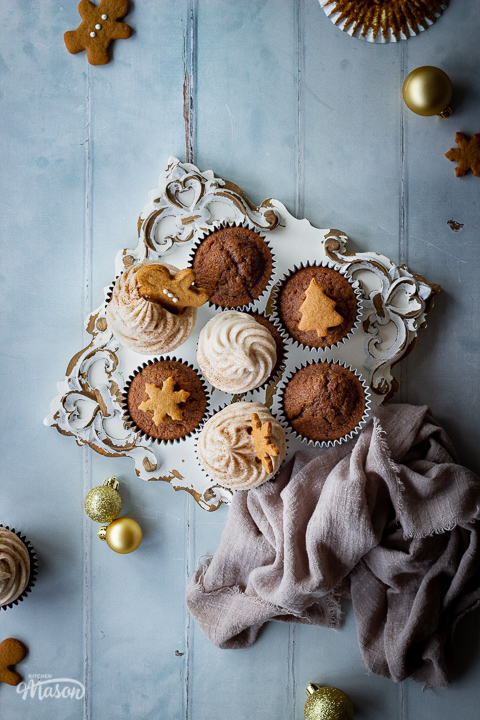 A flat lay view of a mixture of iced and non iced gingerbread cupcakes on a decorative white board. There are mini gingerbread biscuits, gold baubles, more cupcakes and a light brown linen napkin in the background. Set on a cool grey wood effect backdrop.