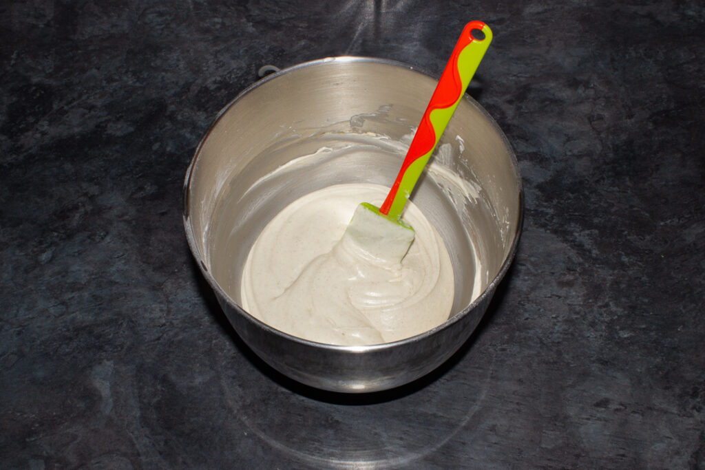 Cinnamon cream cheese frosting in a large silver bowl with a spatula. Set on a kitchen worktop.
