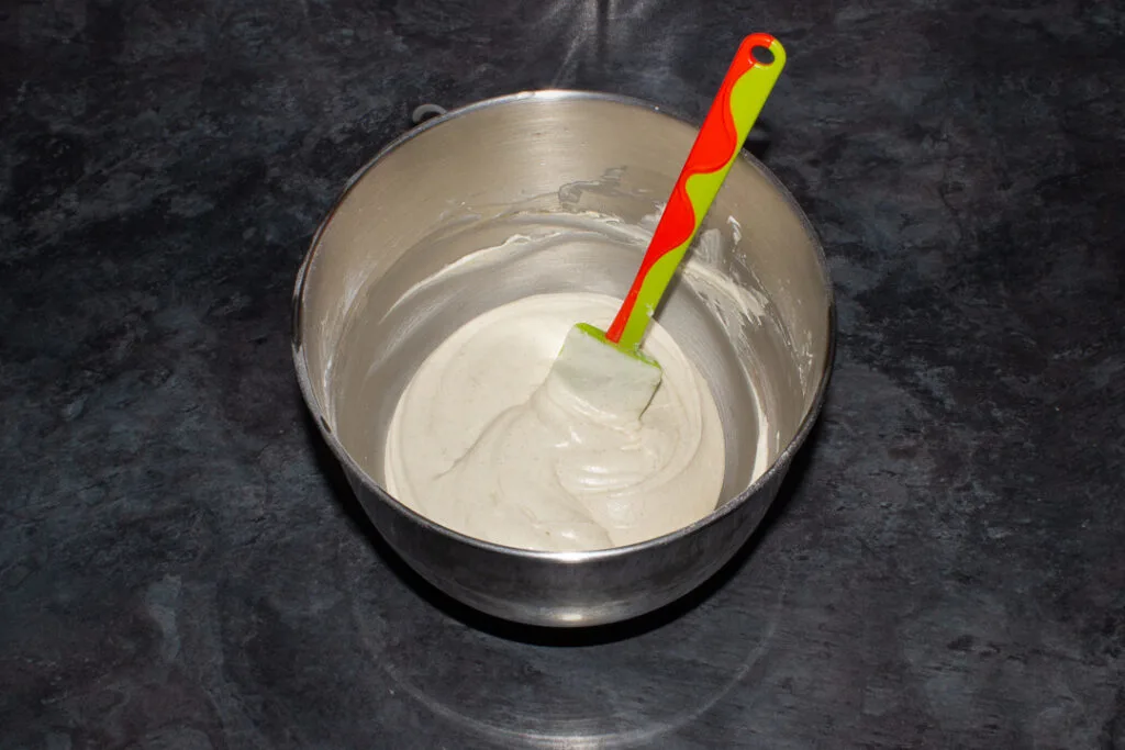 Cinnamon cream cheese frosting in a large silver bowl with a spatula. Set on a kitchen worktop.