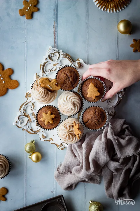 A flat lay view of a hand reaching into a mixture of iced and non iced gingerbread cupcakes on a decorative white board. There are mini gingerbread biscuits, gold baubles, more cupcakes, a muffin pan and a light brown linen napkin in the background. Set on a cool grey wood effect backdrop.