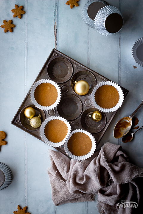A top down view of some unbaked gingerbread cupcakes in a muffin pan with some gold baubles, cupcake cases and mini gingerbread biscuits scattered around in the background with a light brown linen napkin. Set on a cool grey wood effect backdrop.