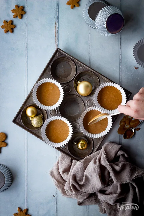 A top down view of a hand reaching in with a spoon to some unbaked gingerbread cupcakes in a muffin pan with some gold baubles, cupcake cases and mini gingerbread biscuits scattered around in the background with a light brown linen napkin. Set on a cool grey wood effect backdrop.