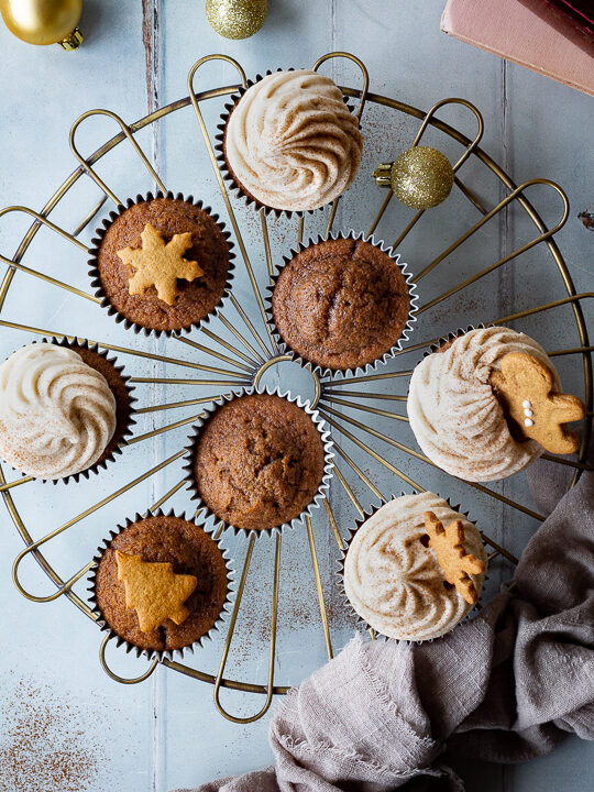 A top down view of a mixture of iced and none iced gingerbread cupcakes on a cooling rack. There is a mini sieve filled with cinnamon, some books, mini gingerbread biscuits, gold baubles and a light brown linen napkin in the background. Set on a cool grey wood effect backdrop.