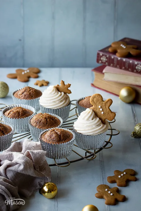 A mixture of iced and none iced gingerbread cupcakes on a round cooling rack. There are some mini gingerbread biscuits, gold baubles, books and a light brown linen napkin in the backlground. Set on a cool grey wood effect backdrop.