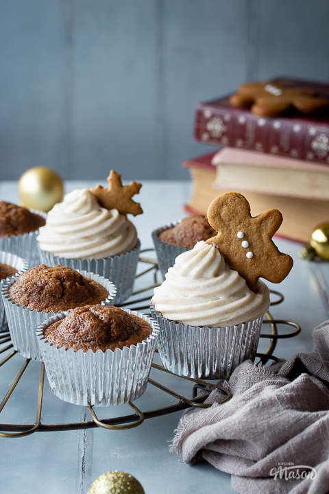 A close up of a mixture of iced and none iced gingerbread cupcakes on a round cooling rack. There are some gold baubles, books and a light brown linen napkin in the background. Set on a cool grey wood effect backdrop.