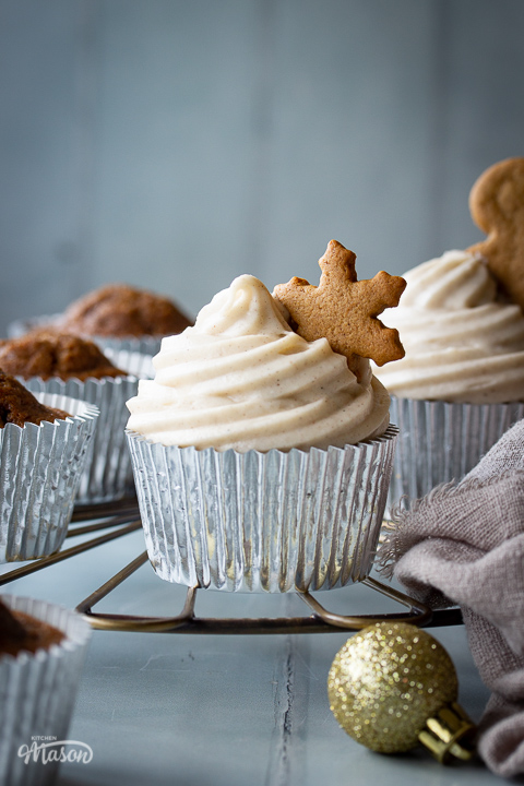A close up of a gingerbread cupcake topped with cream cheese frosting and a gingerbread star. Set on a cooling rack with more cupcakes, gold baubles and a light brown linen napkin in the background. Set on a cool grey wood effect backdrop.