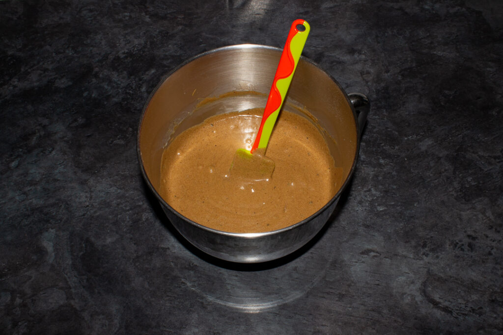 Gingerbread cupcake batter in a large silver bowl with a spatula inside. Set on a kitchen worktop.
