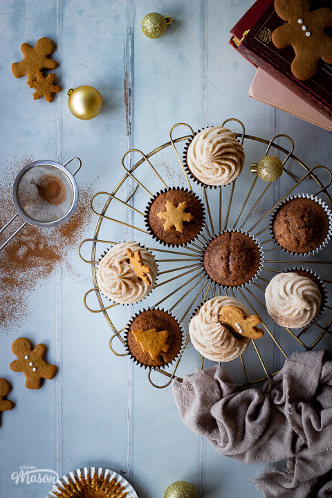 A top down view of a mixture of iced and none iced gingerbread cupcakes on a cooling rack. There is a mini sieve filled with cinnamon, some books, mini gingerbread biscuits, gold baubles and a light brown linen napkin in the background. Set on a cool grey wood effect backdrop.