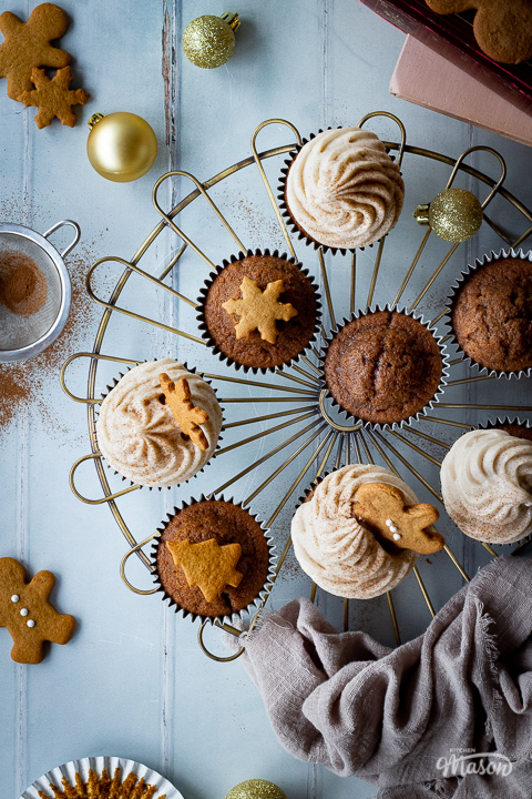 A close up top down view of a mixture of iced and none iced gingerbread cupcakes on a cooling rack. There is a mini sieve filled with cinnamon, some books, mini gingerbread biscuits, gold baubles and a light brown linen napkin in the background. Set on a cool grey wood effect backdrop.